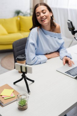 Cheerful and blurred podcaster looking at smartphone on tripod near microphone and using laptop near takeaway coffee during stream in studio  clipart