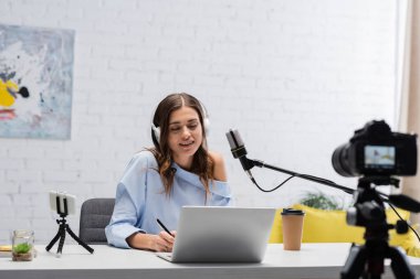 Kulaklıkla gülümseyen esmer podcaster dizüstü bilgisayar kullanıyor ve stüdyoda akış sırasında mikrofonun yanında kalem ve dijital kamera tutuyor. 