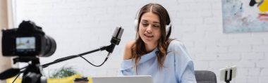 Positive brunette broadcaster in headphones talking during stream near microphone, gadgets and coffee to go next to digital camera in podcast  studio, banner  clipart