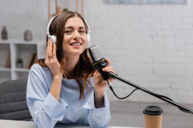 Smiling brunette podcaster in wireless headphones touching microphone and looking at camera near coffee to go in paper cup during stream in studio  clipart