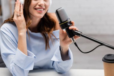 Kablosuz kulaklıklarla mikrofonun yanında konuşan bulanık ve pozitif podcast sunucunun görüntüsü ve stüdyodaki kağıt bardakta kahve içmek. 