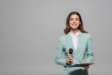 Carefree and brunette event host in blue formal wear holding microphone and clipboard while looking at camera and standing isolated on grey with copy space clipart