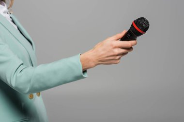 Cropped view of event host in green jacket holding wireless microphone during party and performance while standing isolated on grey   clipart