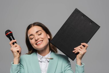 Smiling brunette event host with closed eyes in blue jacket holding microphone and clipboard during holiday celebration isolated on grey   clipart