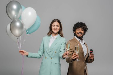 Smiling multiethnic event hosts in formal wear holding festive balloons, microphone and glasses champagne while looking at camera during party isolated on grey   clipart