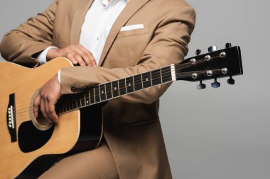 Cropped view of young indian man in brown formal wear holding acoustic guitar during performance while sitting isolated on grey with copy space, in studio  clipart