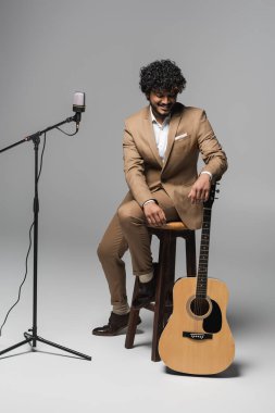 Young and smiling indian event host in suit looking at acoustic guitar while sitting on chair near microphone during performance  on grey background  clipart