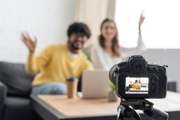 selective focus of professional digital camera near blurred interracial vloggers waving hands near laptop and coffee to go while recording podcast in radio studio