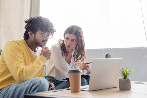 брюнетка женщина указывает на ноутбук во время видеозвонка рядом с серьезным индийским коллегой в очках и желтый свитер рядом с кофе, чтобы пойти и размыть мобильный телефон в студии вещания