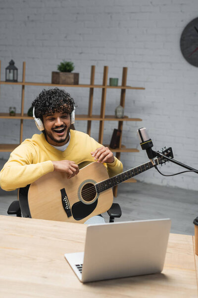 curly indian man in wireless headphones and yellow jumper laughing while sitting with acoustic guitar near laptop and professional microphones in studio
