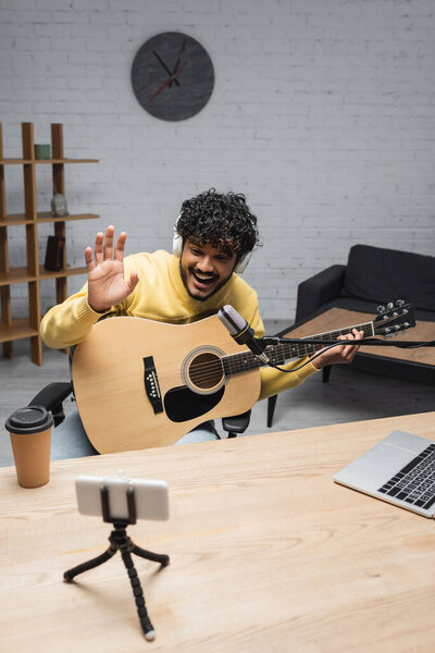 Young and positive indian podcaster in wireless headphones waving hand at smartphone during video call and holding acoustic guitar near coffee to go and laptop on table in podcast studio 
