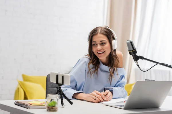 Smiling brunette podcaster in wireless headphones using smartphone on tripod and laptop near microphone and notebooks on table during stream in studio