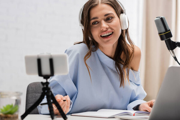 Smiling brunette blogger in wireless headphones talking during stream on blurred smartphone on tripod near microphone and laptop in studio 