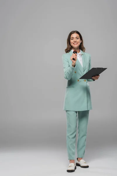 stock image Full length of smiling and brunette event host in blue suit holding microphone and clipboard while looking at camera and standing on grey background 