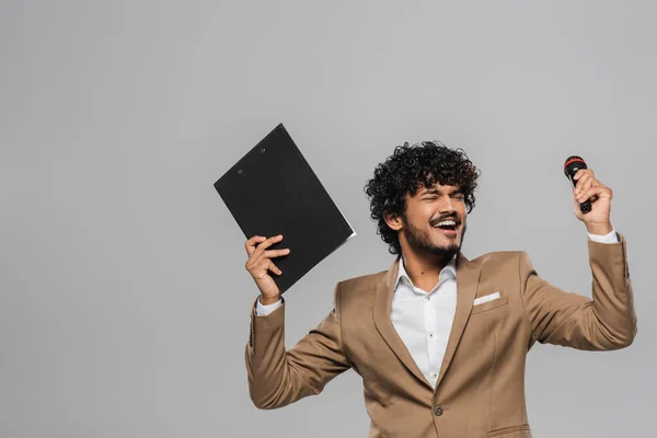 Excited young indian event host with curly hair in formal wear closing eyes while holding clipboard and microphone during performance isolated on grey