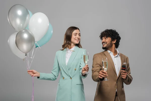 stock image Positive interracial event hosts in formal wear with microphone and festive balloons holding champagne glasses and looking at each other isolated on grey  