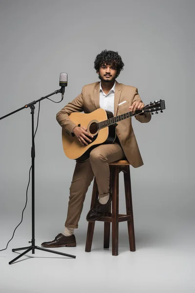 Full length of indian event host in formal wear playing acoustic guitar near microphone during performance while sitting on chair on grey background