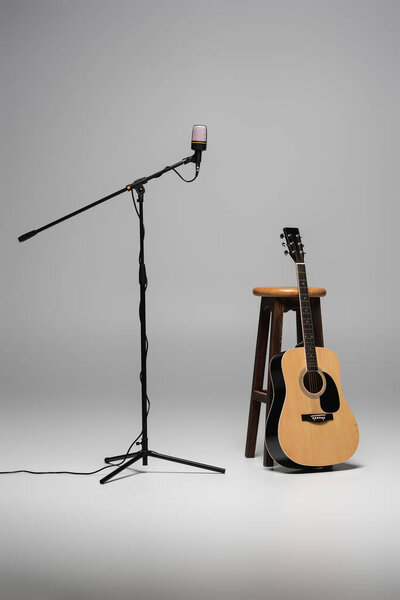 Microphone on metal stand and acoustic guitar near wooden brown chair on grey background 
