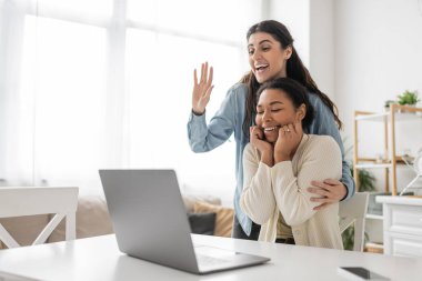 happy lesbian woman waving hand during video call near multiracial girlfriend with engagement ring on finger clipart