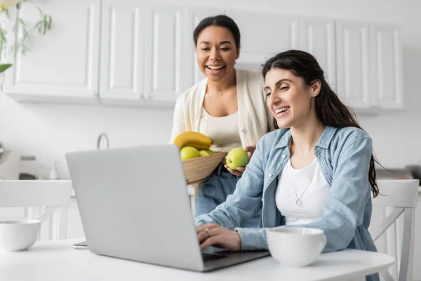 快乐的多种族女人拿着盛有水果的碗 几乎是快乐的女朋友在家里的笔记本电脑上工作 — 图库照片