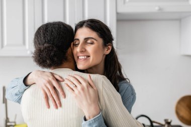 overjoyed lesbian woman with wedding ring on finger smiling and hugging multiracial girlfriend   clipart