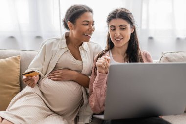 Hamile, çok ırklı bir kadın elinde kredi kartı tutarken neşeli lezbiyen bir partnerle online alışveriş yapıyor. 