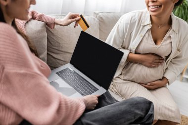 Birlikte online alışveriş yaparken hamile çok ırklı bir ortağın yanında kredi kartı tutan neşeli lezbiyen kadın görüntüsü. 