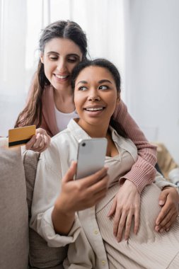 Mutlu ve hamile çok ırklı kadın elinde akıllı telefon tutarken lezbiyen partnerle internetten alışveriş yapıyor. 