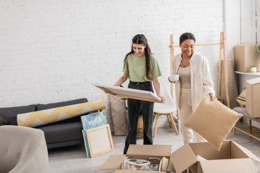 happy multiracial woman holding cup of coffee near cheerful girlfriend with painting in hands  clipart