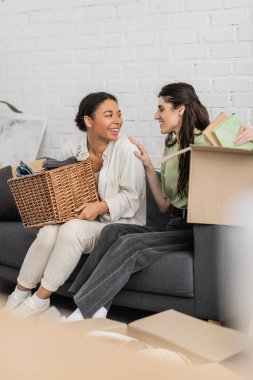 joyful lesbian woman unpacking books from carton box while sitting on sofa next to cheerful multiracial girlfriend   clipart