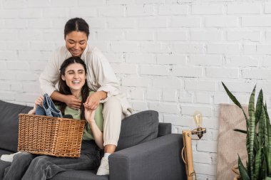 cheerful lesbian woman folding clean laundry while sitting on sofa next to multiracial girlfriend   clipart