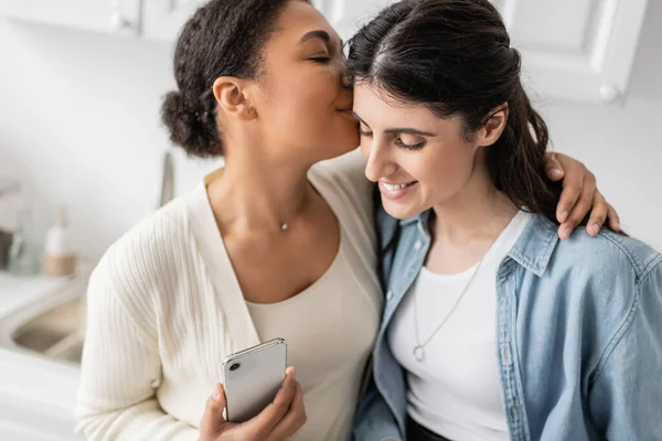 快乐的多种族女性 手持智能手机 亲吻同性恋伴侣的面颊 — 图库照片