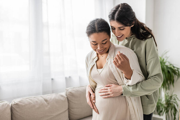 cheerful lesbian woman smiling while hugging belly of pregnant multiracial wife in living room 