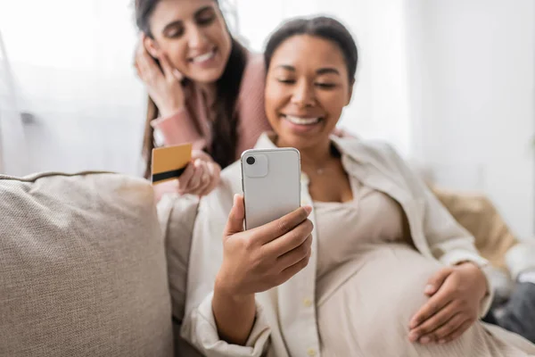幸せなレズビアンパートナーとオンラインショッピングをしながらスマートフォンを見ている陽気で妊娠中の多人種女性 — ストック写真