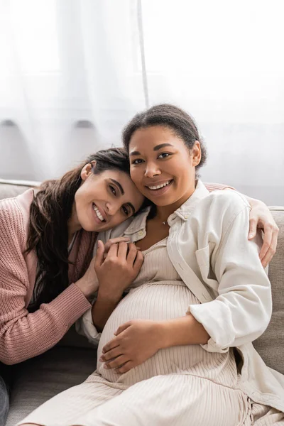 Feliz Lesbiana Mujer Abrazando Positivo Embarazada Multirracial Pareja Sentado Sofá — Foto de Stock