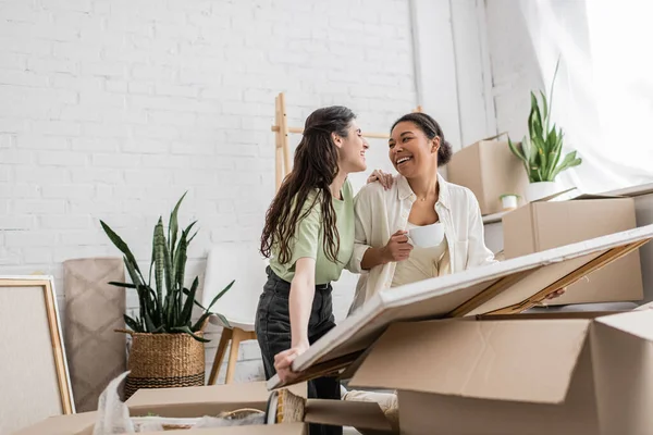 Joyful Interracial Lesbian Couple Looking Each Other While Unpacking Painting — Stock Photo, Image