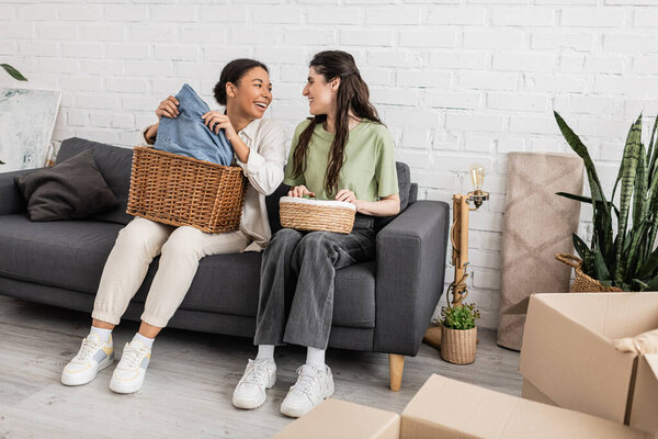joyful interracial lesbian couple holding wicker baskets while sitting on sofa next to plants 