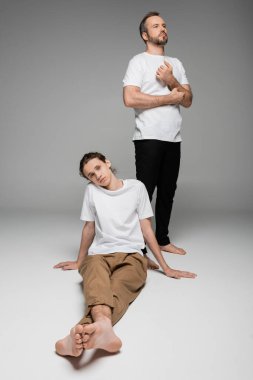 Beyaz tişörtlü, gri tişörtlü bir çocuğun yanında duran uzun boylu bir baba. 