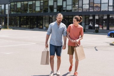 bearded father walking next to happy teenage son and holding shopping bags near mall  clipart
