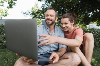 Gülümseyen baba yeşil çimlerde otururken mutlu gencin yanında dizüstü bilgisayar kullanıyor. 