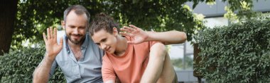 happy father and teenager son waving hands while sitting together in green park, banner  clipart