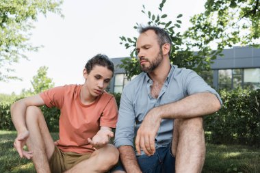 bearded father sitting next to teenage son while talking in green park  clipart