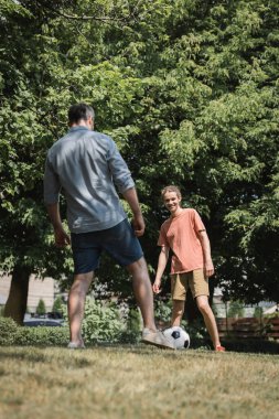 Mutlu genç çocuk babasıyla yeşil yaz parkında futbol oynuyor. 