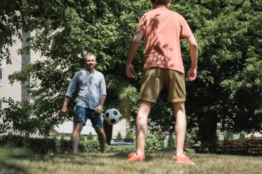 Yeşil yaz parkında oğluyla futbol oynayan sakallı bir adam. 