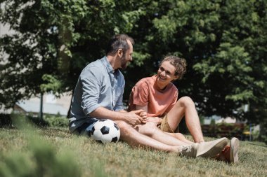 Yeşil yaz parkında babasıyla futbol topunun yanında oturan mutlu genç çocuk. 