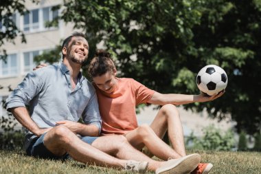 Genç çocuk neşeli babasının yanında oturuyor ve yeşil yaz parkında futbol topu tutuyor. 