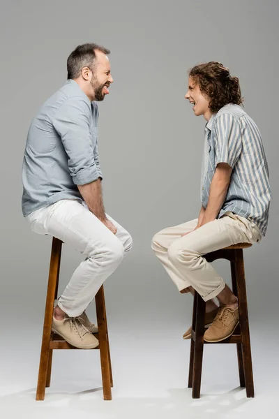 side view of funny dad and teenage son grimacing at each other while sitting on high chairs on grey