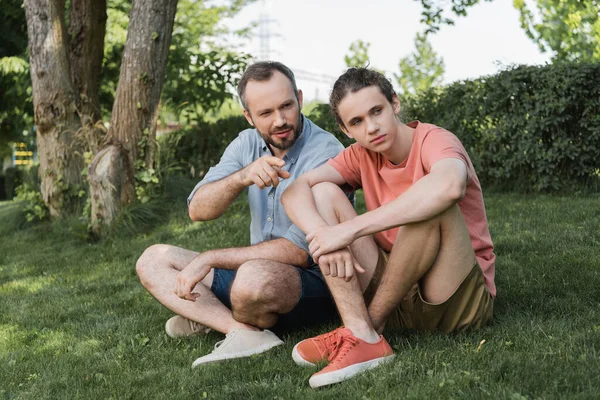满脸胡须的男人一边指指点着近于十几岁的儿子 一边坐在青绿的草坪上 — 图库照片
