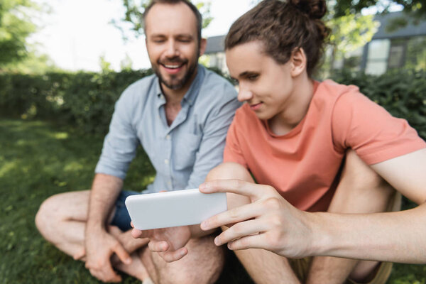 happy teenage boy holding smartphone near bearded father in green park 