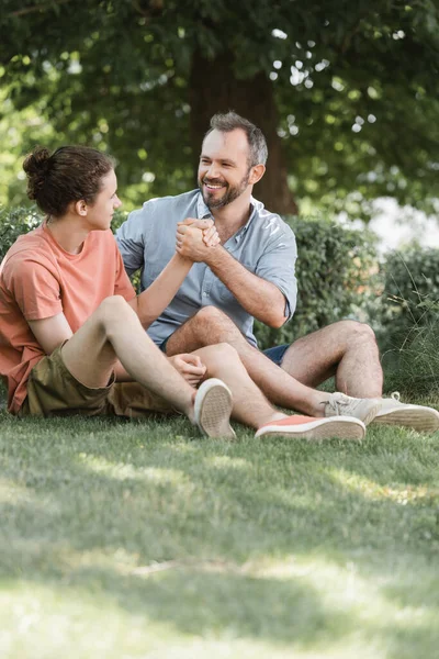 快乐的父亲和儿子坐在公园的绿色草坪上握手 — 图库照片